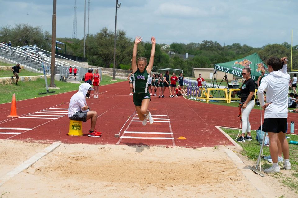 Atleta da SMA no ar durante o salto em distância em uma competição de atletismo em casa, com arquibancada ao fundo