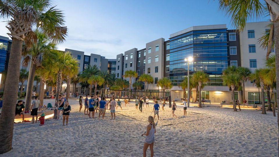 Estudantes jogam vôlei de areia ao anoitecer, com palmeiras e as residências estudantis iluminadas ao fundo, no campus da Embry-Riddle em Daytona Beach