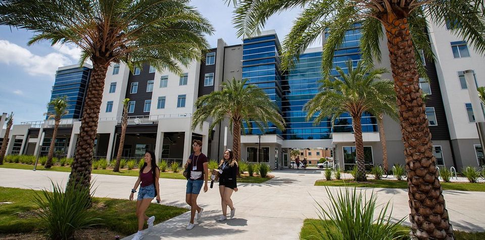 Três estudantes caminham em frente a uma residência estudantil moderna de vidro e concreto no campus da Embry-Riddle em Daytona Beach, entre palmeiras
