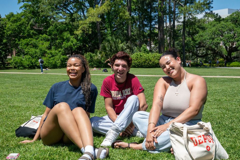 Três estudantes sorrindo sentados na grama do Landis Green da FSU, um deles com camiseta garnet da Florida State, sob o sol de Tallahassee