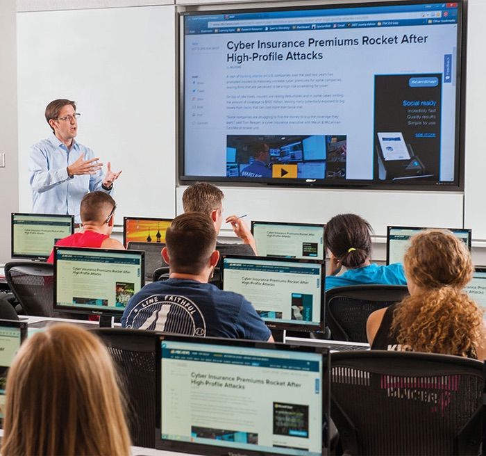 Aula no laboratório de cibersegurança do Innovation and Collaboration Building, com o professor diante de um telão e alunos em computadores