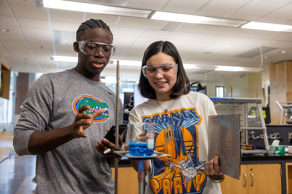 Dois estudantes de óculos de proteção examinam um béquer com líquido azul em um laboratório de química da University of Florida