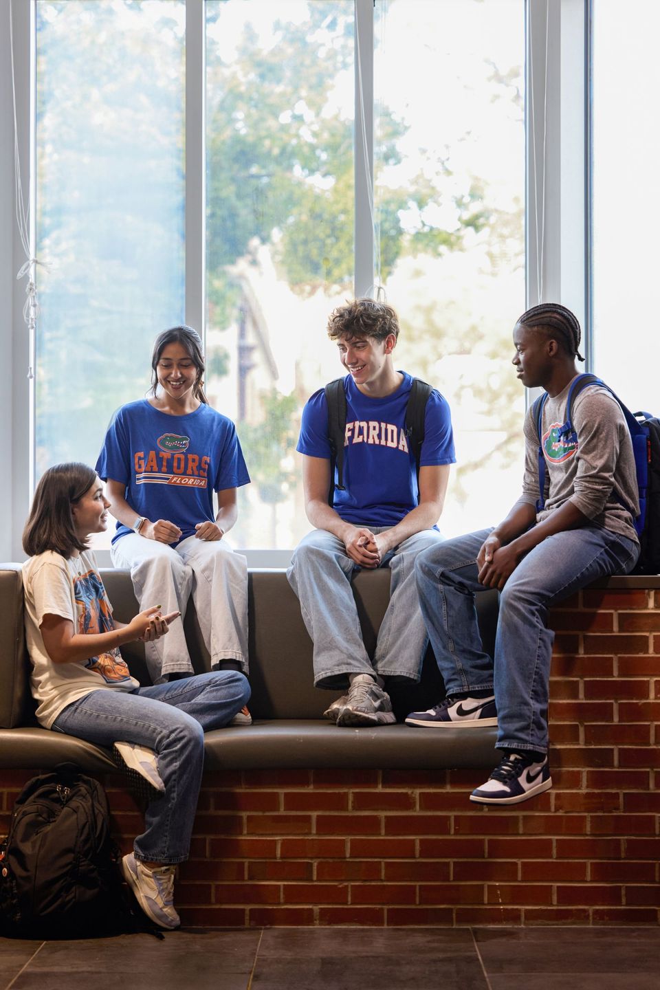 Quatro estudantes com camisetas da Florida e dos Gators conversam sentados em um banco de tijolos diante de uma janela ampla, em uma área comum do campus da University of Florida